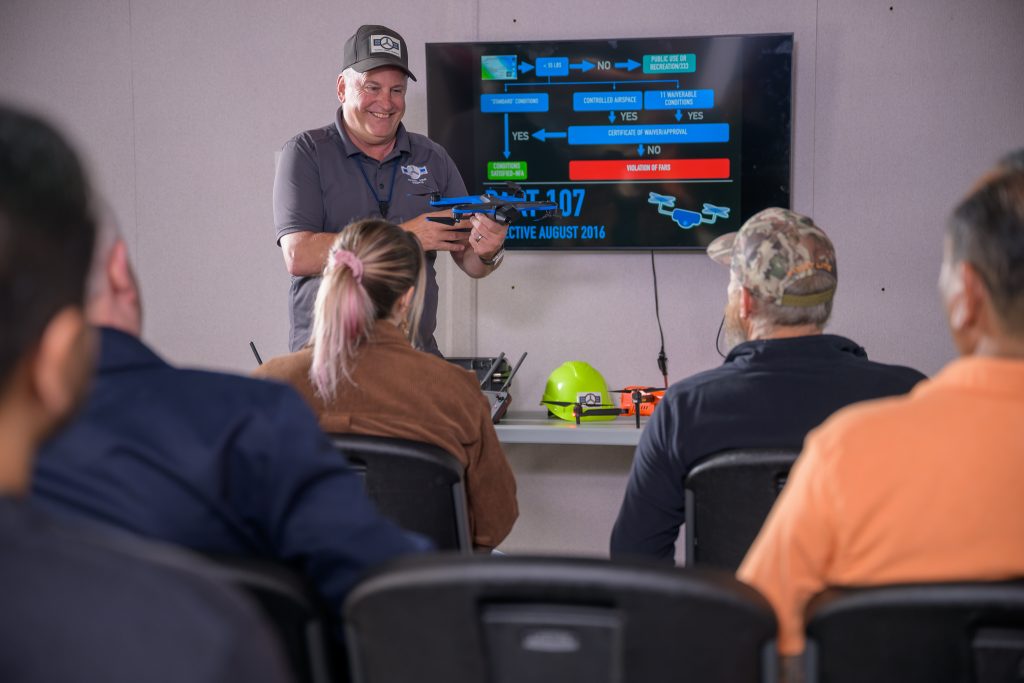 Male instructor standing infront of a classroom of adult students holding a drone